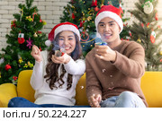 Young asian adult teenager couple celebrateing christmas holiday together with Firecrackers in living room with christmas tree decoration. Стоковое фото, агентство Ingram Publishing / Фотобанк Лори