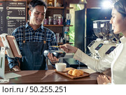 Asian customer using her credit card with contactless nfs technology to pay a barista for her coffee purchase at a cafe bar. Стоковое фото, агентство Ingram Publishing / Фотобанк Лори