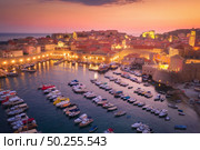 Aerial view of boats and yachts and beautiful architecture at night in Dubrovnik, Croatia. Top view of old city at sunset. Port in the sea, city lights, historical centre, buildings and colorful sky. Стоковое фото, агентство Ingram Publishing / Фотобанк Лори