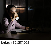Young adult asian businesswoman drinking hot cofee while working late at night in her office with desktop computer. Using as hard working and working late concept. Стоковое фото, агентство Ingram Publishing / Фотобанк Лори
