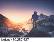 Man is standing on the mountain peak against snowy mountains, fjord at colorful sunset in winter. Young man on the stone, sea coast and rocks, blue sky in Lofoten Islands, Norway. Travel and hiking. Стоковое фото, агентство Ingram Publishing / Фотобанк Лори