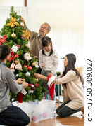Multigenerational asian Family decorating a Christmas tree. Mom Dad daughter girl and grandfather decorate the Christmas tree prepare for season greeting of Merry Christmas and Happy Holidays. Стоковое фото, агентство Ingram Publishing / Фотобанк Лори