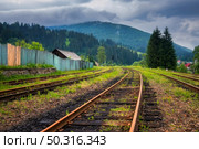 Railroad in mountains in overcast summer day. Industrial landscape with old railway station, trees, green grass, buildings, hills in fog, moody sky with clouds in fall. Railway platform in village. Стоковое фото, агентство Ingram Publishing / Фотобанк Лори