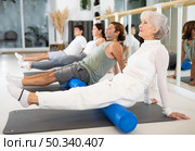 Elderly woman practicing pilates with roller on gray mat in gym room. Стоковое фото, фотограф Яков Филимонов / Фотобанк Лори