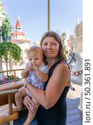 A mother smiles while cradling her baby, both enjoying a bright day at a vibrant theme park filled with attractions. Стоковое фото, фотограф Данила Большаков / Фотобанк Лори