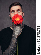 contemplated young man with pierced nose ears holding red gerbera flower front his mouth against grey background. Стоковое фото, агентство Ingram Publishing / Фотобанк Лори