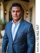 Confident man in a blue suit is standing in front of a house, looking towards the camera. The house has a white picket fence and green lawn. Стоковое AI-изображение, AI-художник Данила Большаков / Фотобанк Лори