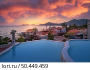 Beautiful swimming pool, sky with pink clouds at colorful sunset. Blue water, sea coast, architecture, palm trees in summer. Luxury resort. Oludeniz, Turkey. Landscape with pool, houses, orange roofs. Стоковое фото, агентство Ingram Publishing / Фотобанк Лори