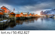 Beautiful orange rorbu in overcast sunset. Fishing village in Lofoten islands, Norway. Winter landscape with houses, snowy mountains, blue sea, cloudy sky. Norwegian traditional rorbuer on the water. Стоковое фото, агентство Ingram Publishing / Фотобанк Лори