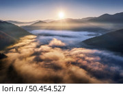 Mountains in low clouds at sunrise in autumn. Aerial view of mountain peak in fog in fall. Beautiful landscape with rocks, forest, sun, purple sky. Top view of mountain valley in clouds. Foggy hills. Стоковое фото, агентство Ingram Publishing / Фотобанк Лори
