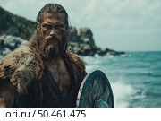 Atmospheric portrait of a Viking against the backdrop of a rugged northern coast. A masculine bearded warrior in furs and leather armor holds a wooden shield, his face covered in dirt and battle marks. His gaze is purposeful and determined, his hair is tousled by the wind. Behind him is a raging sea and a rocky shore, emphasizing the strength and spirit of the warrior. The image is ideal for the themes of history, mythology, fantasy, military affairs, reconstruction and epic narratives. Стоковое AI-изображение, AI-художник Анна Кривицкая / Фотобанк Лори