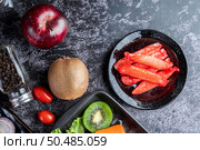 Crab stick put on a black plate with raw salmon, tomatoes, kiwi, shallots and sweet peppers on the black cement floor. Стоковое фото, агентство Ingram Publishing / Фотобанк Лори