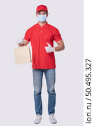 Image of a happy young delivery man in red cap blank t-shirt uniform face mask gloves standing with empty brown craft paper packet isolated on light gray background studio, Service during pandemic coronavirus virus covid-19 concept. Стоковое фото, агентство Ingram Publishing / Фотобанк Лори