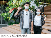 Cheerful Young couple in mask holding multiple paper shopping bag going down stairs of large shopping mall and pretty woman show credit card in hand,  Lifestyle and shopping concept, copy space. Стоковое фото, агентство Ingram Publishing / Фотобанк Лори