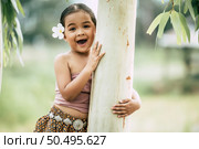 Close up, Portrait of little girl  in Thai traditional dress and put white flower on her ear, Stand and embrace trunk of the tree, laugh and looking at camera, copy space. Стоковое фото, агентство Ingram Publishing / Фотобанк Лори