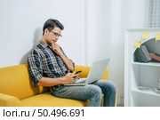 Young handsome man wearing eyeglasses with new normal, sitting on sofa and works from home on laptop computer while self-isolation and quarantine covid-19 coronavirus outbreak. Стоковое фото, агентство Ingram Publishing / Фотобанк Лори