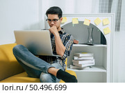 Young handsome man wearing eyeglasses with new normal, sitting on sofa and works from home on laptop computer while self-isolation and quarantine covid-19 coronavirus outbreak. Стоковое фото, агентство Ingram Publishing / Фотобанк Лори