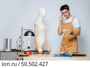 Portrait Young male fashion designer use smartphone during working with mannequin and take a note at the studio at home,  full of tailoring tools and equipment. Стоковое фото, агентство Ingram Publishing / Фотобанк Лори