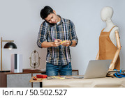 young Asian fashion designer or tailor working with laptop computer receive orders on smartphone, full of tailoring tools with mannequin and equipment on desk in studio. Стоковое фото, агентство Ingram Publishing / Фотобанк Лори