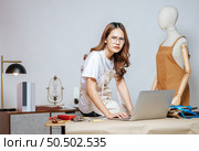 Seriously young Asian fashion designer or tailor working with laptop computer to search idea or online orders, full of tailoring tools  with mannequin and equipment on the desk in the studio. Стоковое фото, агентство Ingram Publishing / Фотобанк Лори