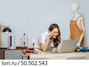 Beautiful young Asian fashion designer or tailor working with laptop computer to search idea, inspiration or online orders, full of tailoring tools with mannequin and equipment on desk in studio. Стоковое фото, агентство Ingram Publishing / Фотобанк Лори
