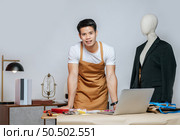 Portrait Young handsome male designer and laptop computer for searching good idea to design fashion clothing, mannequin and equipment with full of tailoring tools on desk in studio. Стоковое фото, агентство Ingram Publishing / Фотобанк Лори