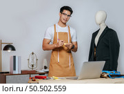Cheerful young Asian fashion designer or tailor wearing eyeglasses use laptop computer to search idea or online orders, mannequin in suit and full of tailoring tools with equipment on desk in studio. Стоковое фото, агентство Ingram Publishing / Фотобанк Лори