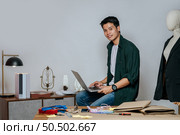 Portrait Young handsome male designer use laptop computer for searching good idea to design fashion clothing, mannequin and equipment with full of tailoring tools on desk in studio. Стоковое фото, агентство Ingram Publishing / Фотобанк Лори