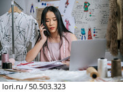Seriously Young designer talking with smartphone while use laptop computer to checking together, full of sewing tool on table, beautiful clothes on hanger. Стоковое фото, агентство Ingram Publishing / Фотобанк Лори