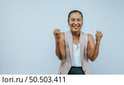 Emotive excited Asian woman keeps hands clenched in fists, exclaims with positiveness as achieved success in her career, smiling widely, isolated over  background in studio. Стоковое фото, агентство Ingram Publishing / Фотобанк Лори
