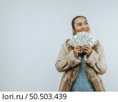 Portrait of successful Asian woman with a fur coat holding money in hand ,smiling and looking aside, standing over background in studio with copy space. Advertising concept. Стоковое фото, агентство Ingram Publishing / Фотобанк Лори