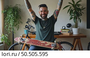 Tattooed lucky freelancer in front of his working space, surrounded with his hobby toys longboard, vintage bicycle and green plant, stretching his hand in air. Стоковое AI-видео, аниматор Sergey Grishin / Фотобанк Лори
