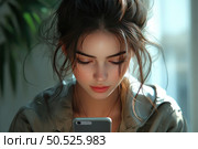 A young woman with natural beauty and expressive features looks intently at the screen of her smartphone. Her face is illuminated by soft daylight, creating an atmosphere of coziness and silence. Her hair is casually gathered in a bun, individual strands freely frame her face. She is wearing casual clothes in neutral tones. The image embodies modernity, concentration and digital reality. Suitable for topics related to technology, communication, mobile devices and youth life. Стоковое AI-изображение, AI-художник Анна Кривицкая / Фотобанк Лори