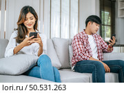 Selective focus, Young Asian Couple sitting on sofa in living room, They are enjoy with social media networks addicts surf the internet on mobile phones ignoring each other, family addicted smartphone concept. Стоковое фото, агентство Ingram Publishing / Фотобанк Лори