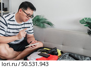 Asian man glasses is preparing clothes in suitcases. He’s choosing clothes, travel documents, the itinerary for a solo trip, and check the checklist. Travel, holiday and vacation concept. Стоковое фото, агентство Ingram Publishing / Фотобанк Лори