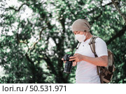 Asian Traveler man wears medical mask with backpack checking a picture on camera at the park with copy space. Travel photographer. Vocation and holiday concept. Стоковое фото, агентство Ingram Publishing / Фотобанк Лори