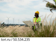 Front view of an engineer wearing a safety vest and a yellow helmet while standing in a field with wind turbines in the background. Стоковое фото, агентство Ingram Publishing / Фотобанк Лори