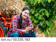 Asian young female farmer sitting on bale of hay with big tractor machine in farmland. Modern technologies in agriculture management and agribusiness concept. Стоковое фото, агентство Ingram Publishing / Фотобанк Лори