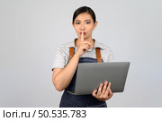 Portrait of young asian woman in waitress uniform holding a laptop computer and finger on mouth posture to make a quiet, copy space to insert your advertisement isolated on white background. Стоковое фото, агентство Ingram Publishing / Фотобанк Лори