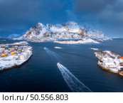 Aerial view of floating fishing boat in blue sea in snowy Henningsvaer village, Lofoten islands, Norway in winter. Top drone view of boat, mountains in snow, sea, cloudy sky, town, rorbuer and houses. Стоковое фото, агентство Ingram Publishing / Фотобанк Лори