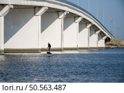 Hydrofoil surfer glides beneath massive concrete bridge, juxtaposing modern water sport technology with urban infrastructure in tranquil waters. Стоковое фото, агентство Ingram Publishing / Фотобанк Лори