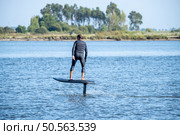 A man in a wetsuit rides a hydrofoil surfboard across calm waters, with trees lining the distant shore on a clear day. Стоковое фото, агентство Ingram Publishing / Фотобанк Лори