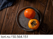 Two ripe persimmons on a round metal plate, with a rustic wooden background and dried leaves. Стоковое фото, агентство Ingram Publishing / Фотобанк Лори