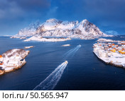 Aerial view of floating fishing boat in blue sea in snowy Henningsvaer village, Lofoten islands, Norway in winter. Top drone view of boat, mountains in snow, sea, cloudy sky, town, rorbuer and houses. Стоковое фото, агентство Ingram Publishing / Фотобанк Лори