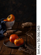 Golden persimmons rest on navy linen against weathered wood. A ceramic bowl and rustic elements create a moody, autumnal scene rich in texture and warmth. Стоковое фото, агентство Ingram Publishing / Фотобанк Лори