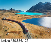 Aerial view of modern red train near azure mountain Bianco lake, snowy rocks and blue sky at sunset in autumn. Bernina Express, Switzerland. Top view of train in Alps, railroad, orange hills in fall. Стоковое фото, агентство Ingram Publishing / Фотобанк Лори