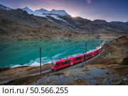 Moving modern red train near azure mountain Bianco lake, snowy rocks and colorful sky at sunset in autumn. Bernina Express, Switzerland. Blurred train in Alps, reflection in water in fall at dusk. Стоковое фото, агентство Ingram Publishing / Фотобанк Лори