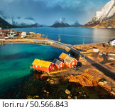 Aerial view of red rorbuer houses, blue sea and snowy mountains at sunset. Winter in Lofoten islands, Norway. Golden sunlight illuminates the vibrant red rorbu cabins of Hamnoy. Top drone view. Travel. Стоковое фото, агентство Ingram Publishing / Фотобанк Лори