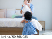 A small girl stood at the foot of the bed playing with the younger sibling carried by his mother and smiling with affection, In a bedroom surrounded by warmth and family. Стоковое фото, агентство Ingram Publishing / Фотобанк Лори
