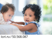 A small girl stood at the foot of the bed playing with the younger sibling carried by his mother and smiling with affection, In a bedroom surrounded by warmth and family. Стоковое фото, агентство Ingram Publishing / Фотобанк Лори